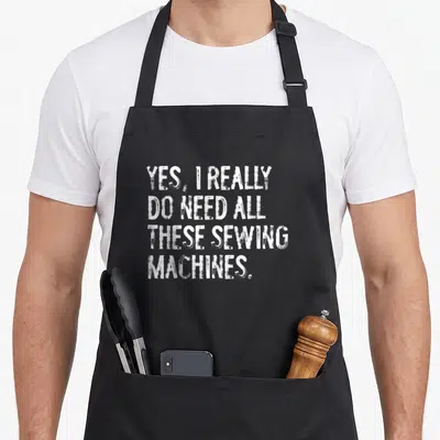 These sewing do i need an apron - White distressed stacked text reading Yes I Really Do Need All These Sewing Machines — shown on a black cotton canvas apron against a white background