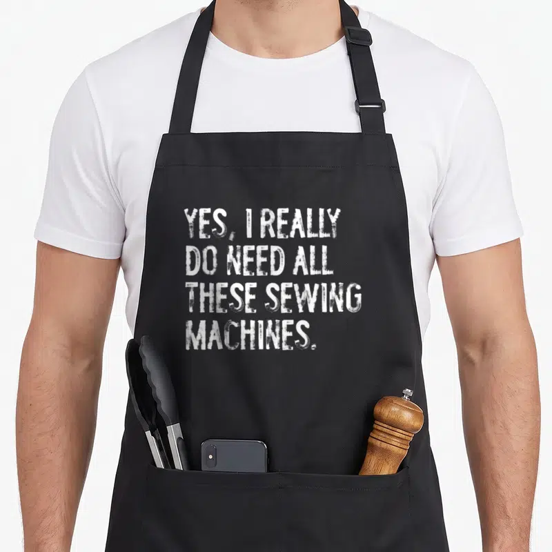 These sewing do i need an apron - White distressed stacked text reading Yes I Really Do Need All These Sewing Machines — shown on a black cotton canvas apron against a white background