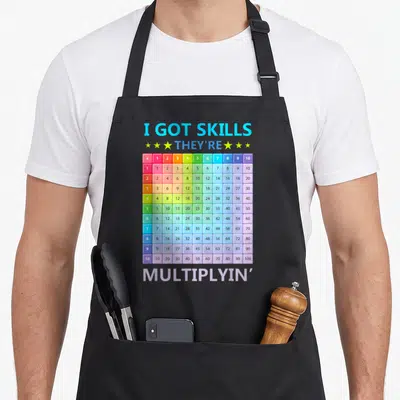 Skill multiplying teacher apron - I GOT SKILLS THEY'RE MULTIPLYIN' text with rainbow multiplication table grid — shown on a black cotton canvas apron against a white background