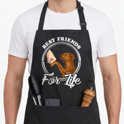 Cool rhodesian ridgeback apron - Brown dog fist bumping human hand in white circle with best friends for life text — shown on a black cotton canvas apron against a white background