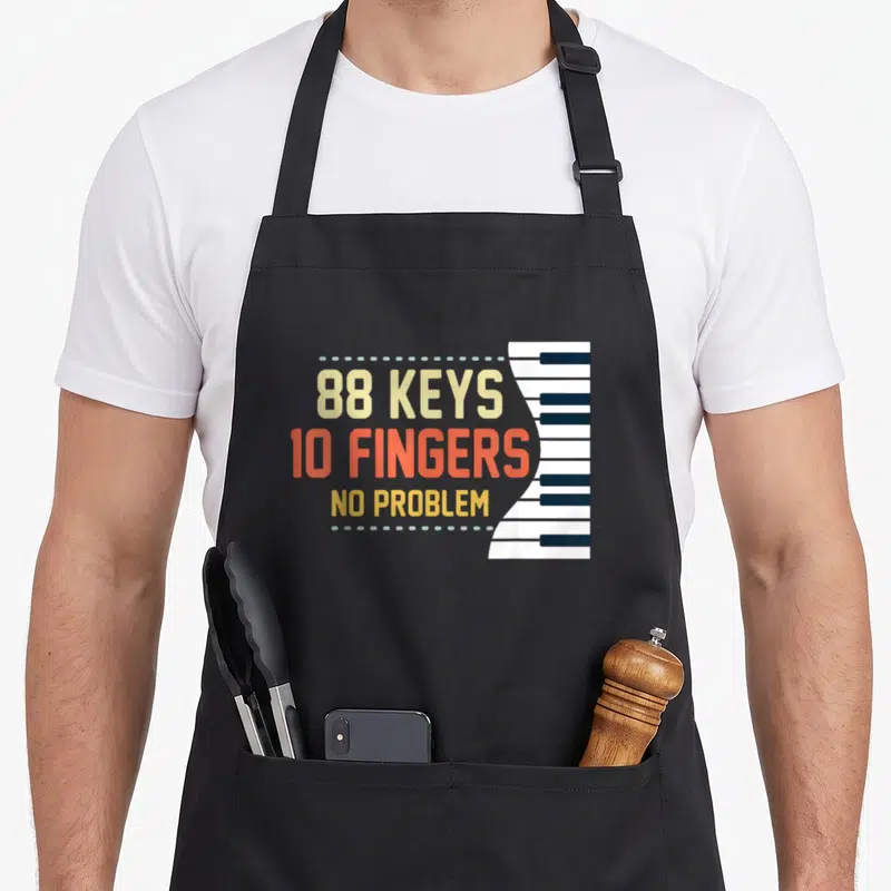 Key musician piano apron - 88 keys 10 fingers no problem text beside vertical piano keyboard in beige and orange-red — shown on a black cotton canvas apron against a white background