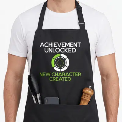 Created character aprons - Baby face inside loading circle with white and lime green text reading Achievement Unlocked — shown on a black cotton canvas apron against a white background