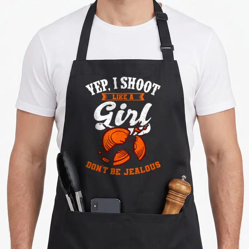 Yep clay apron - Shattered orange clay target with white text YEP I SHOOT LIKE A GIRL — shown on a black cotton canvas apron against a white background
