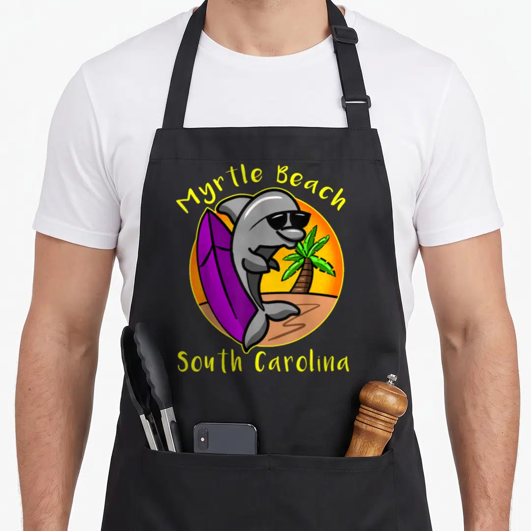 Carolina south beach apron - Grey cartoon dolphin with sunglasses and purple surfboard inside a sunset circle — shown on a black cotton canvas apron against a white background