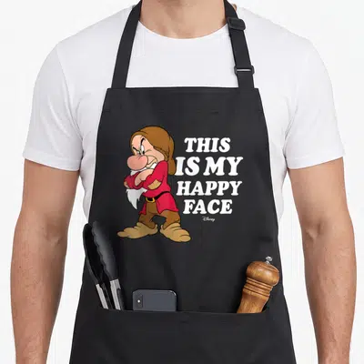 Grumpy princess apron - Cartoon dwarf with crossed arms next to stacked white text reading THIS IS MY HAPPY FACE — shown on a black cotton canvas apron against a white background