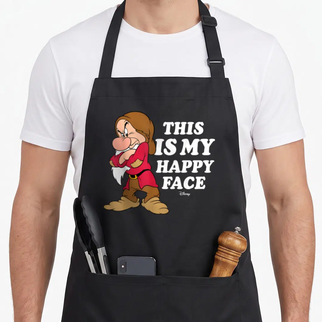 Grumpy princess apron - Cartoon dwarf with crossed arms next to stacked white text reading THIS IS MY HAPPY FACE — shown on a black cotton canvas apron against a white background