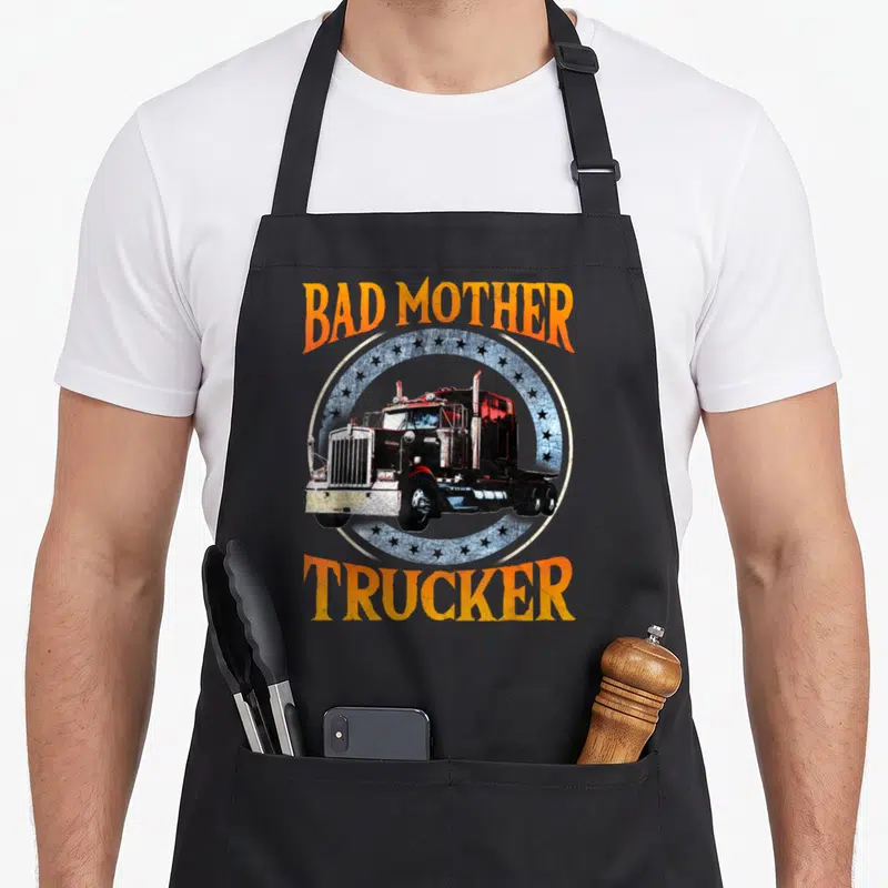 Truck tractor trailer apron - Arched orange text Bad Mother Trucker around a distressed black semi-truck on a blue star — shown on a black cotton canvas apron against a white background