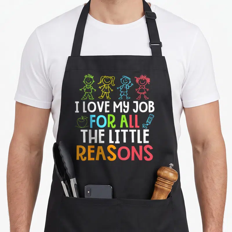 Cute reason teacher apron - Four cartoon children above I LOVE MY JOB FOR ALL THE LITTLE REASONS text with apple — shown on a black cotton canvas apron against a white background
