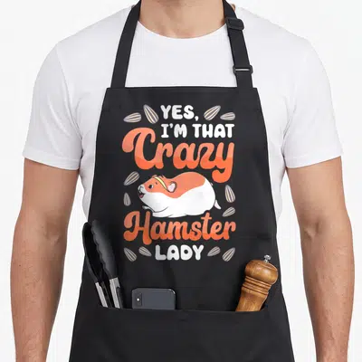 Vintage comfort crazy hamster lady apron - Cartoon hamster with orange script and scattered seeds reading Yes I'm That — shown on a black cotton canvas apron against a white background