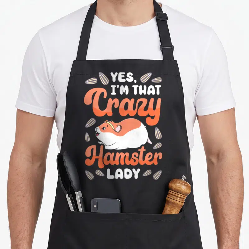Vintage comfort crazy hamster lady apron - Cartoon hamster with orange script and scattered seeds reading Yes I'm That — shown on a black cotton canvas apron against a white background