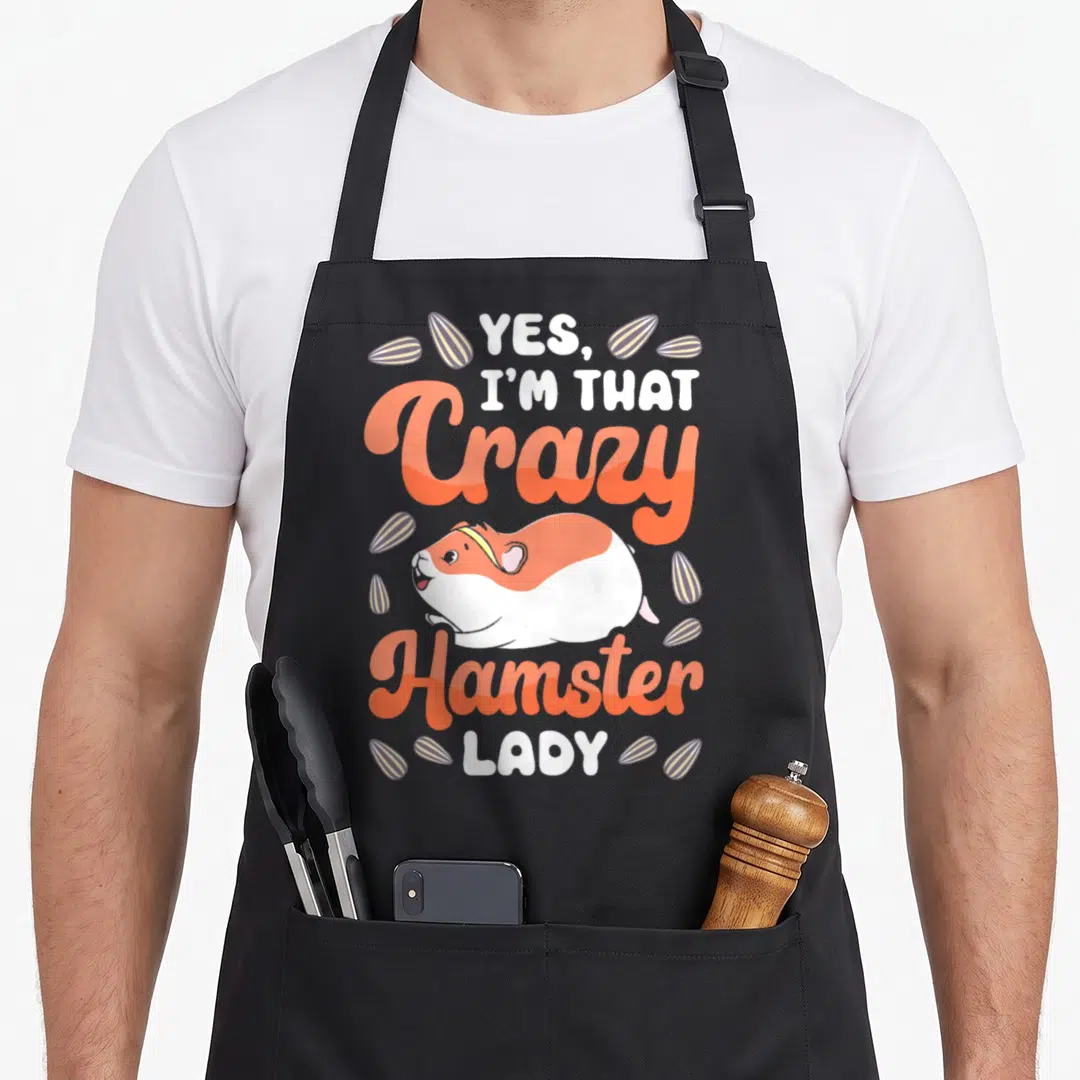 Vintage comfort crazy hamster lady apron - Cartoon hamster with orange script and scattered seeds reading Yes I'm That — shown on a black cotton canvas apron against a white background