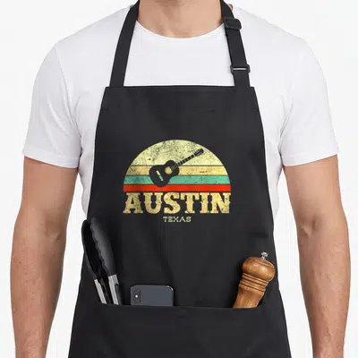 Guitar texas aprons - Retro guitar over yellow orange red and teal stripes with Austin Texas text — shown on a black cotton canvas apron against a white background