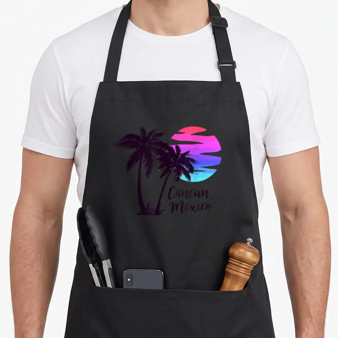 Tree mexico apron - Two dark purple palm trees beside a striped pink and blue gradient sun with Cancun Mexico text — shown on a black cotton canvas apron against a white background