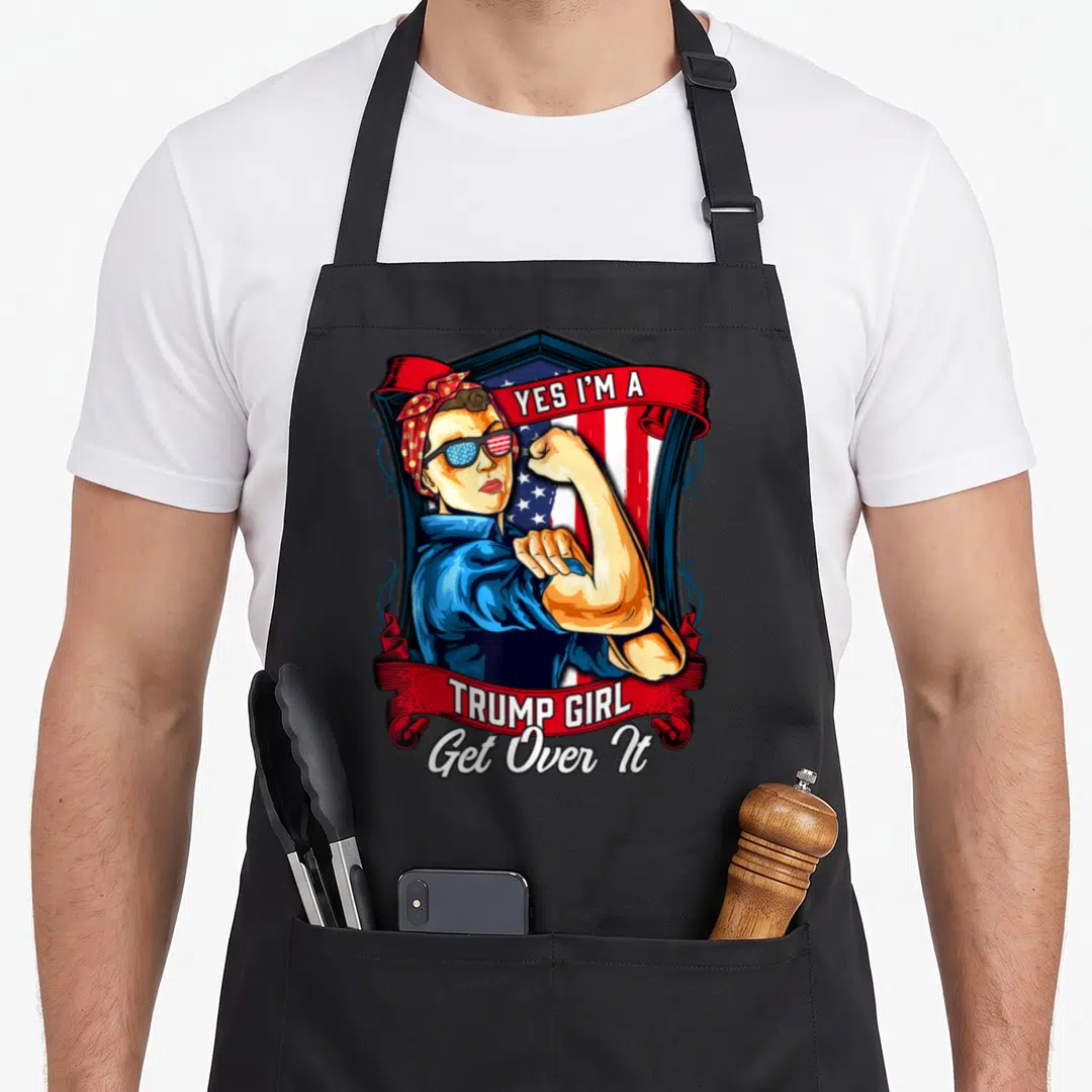 Trump us aprons - Woman flexing arm with Yes I'm A Trump Girl Get Over It text and US flag shield — shown on a black cotton canvas apron against a white background