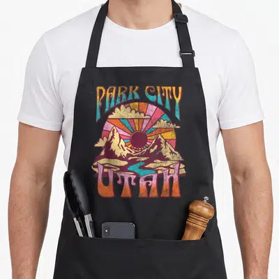 Utah outdoor apron - Retro Park City Utah mountain sunset design with teal river and arched lettering — shown on a black cotton canvas apron against a white background