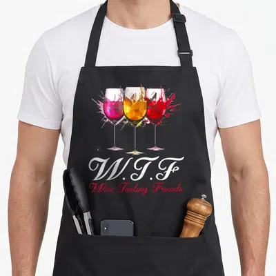 Friend wine apron - Three splashing wine glasses in pink yellow red above W.T.F Wine Tasting Friends text — shown on a black cotton canvas apron against a white background