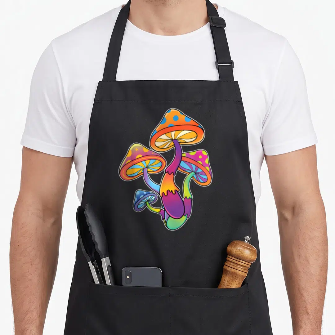 Colorful foraging apron - Four cartoon mushrooms in orange, blue, purple, green, and pink clustered together — shown on a black cotton canvas apron against a white background