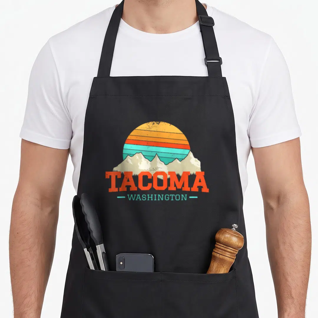 Souvenir tacoma apron - TACOMA WASHINGTON text over beige mountains and striped orange yellow blue sun in retro style — shown on a black cotton canvas apron against a white background