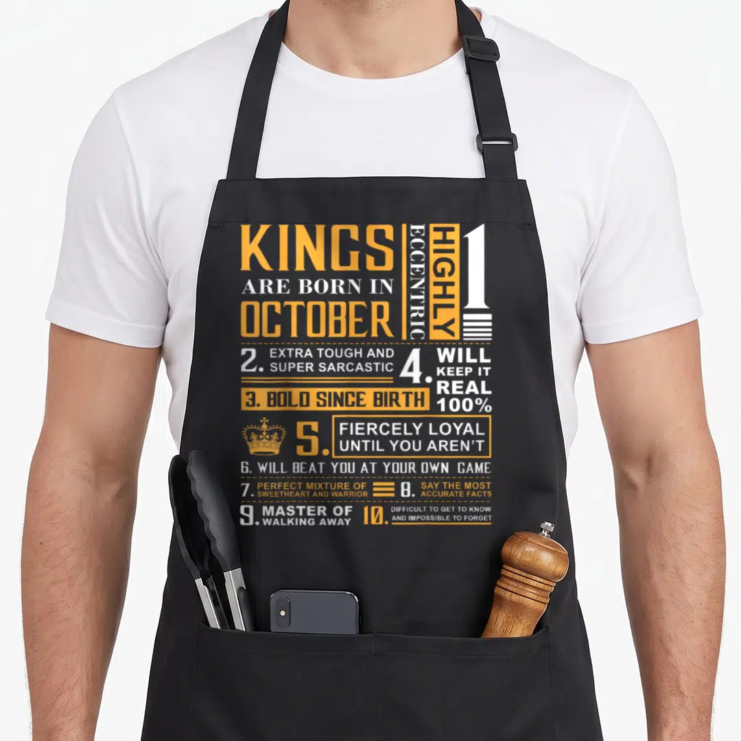 Birthday king apron - Orange and white text grid listing ten traits for Kings born in October with a small crown icon — shown on a black cotton canvas apron against a white background