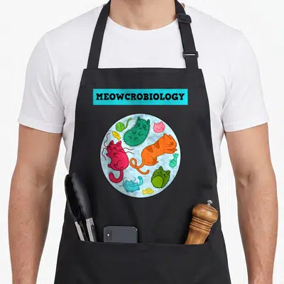 Meowcrobiology science apron - Cartoon cats and fish inside a circle under cyan MEOWCROBIOLOGY text — shown on a black cotton canvas apron against a white background