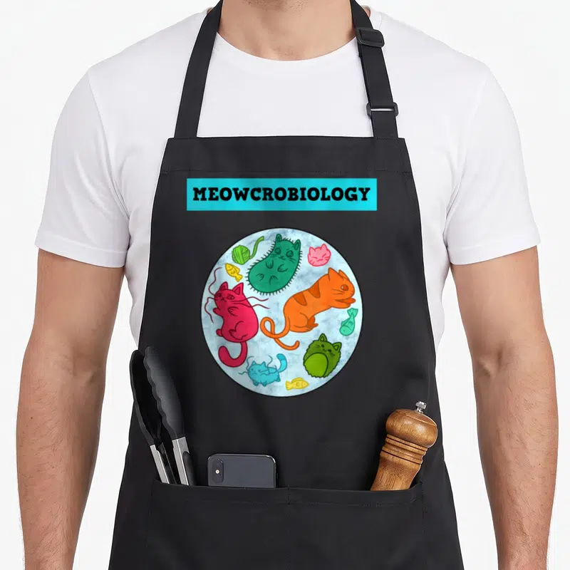 Meowcrobiology science apron - Cartoon cats and fish inside a circle under cyan MEOWCROBIOLOGY text — shown on a black cotton canvas apron against a white background