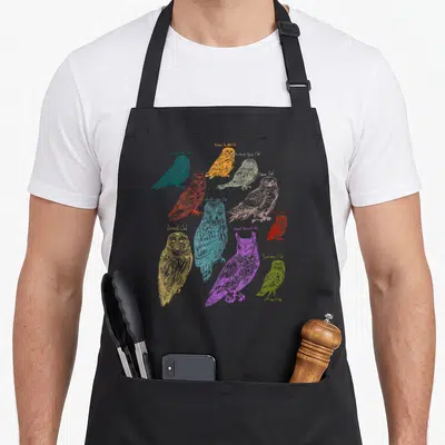 Forest owl apron - Nine colored line-art owls labeled with species names like Barn Owl and Snow Owl — shown on a black cotton canvas apron against a white background