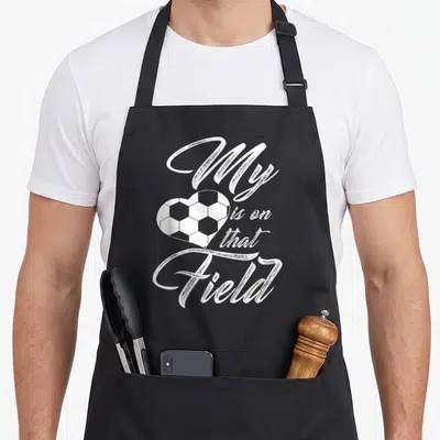 Field funny soccer apron - White heart shaped soccer ball graphic with My is on that Field text — shown on a black cotton canvas apron against a white background