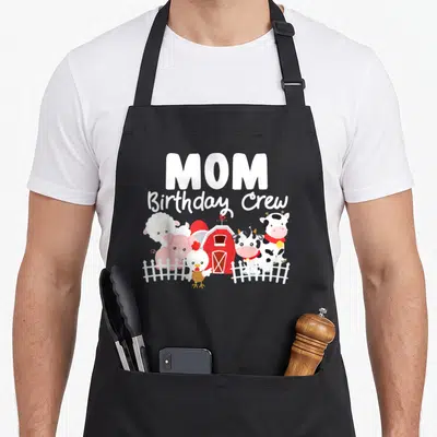 Birthday farm crew aprons - MOM Birthday Crew text above cartoon cow pig sheep chicken and red barn behind white fence — shown on a black cotton canvas apron against a white background