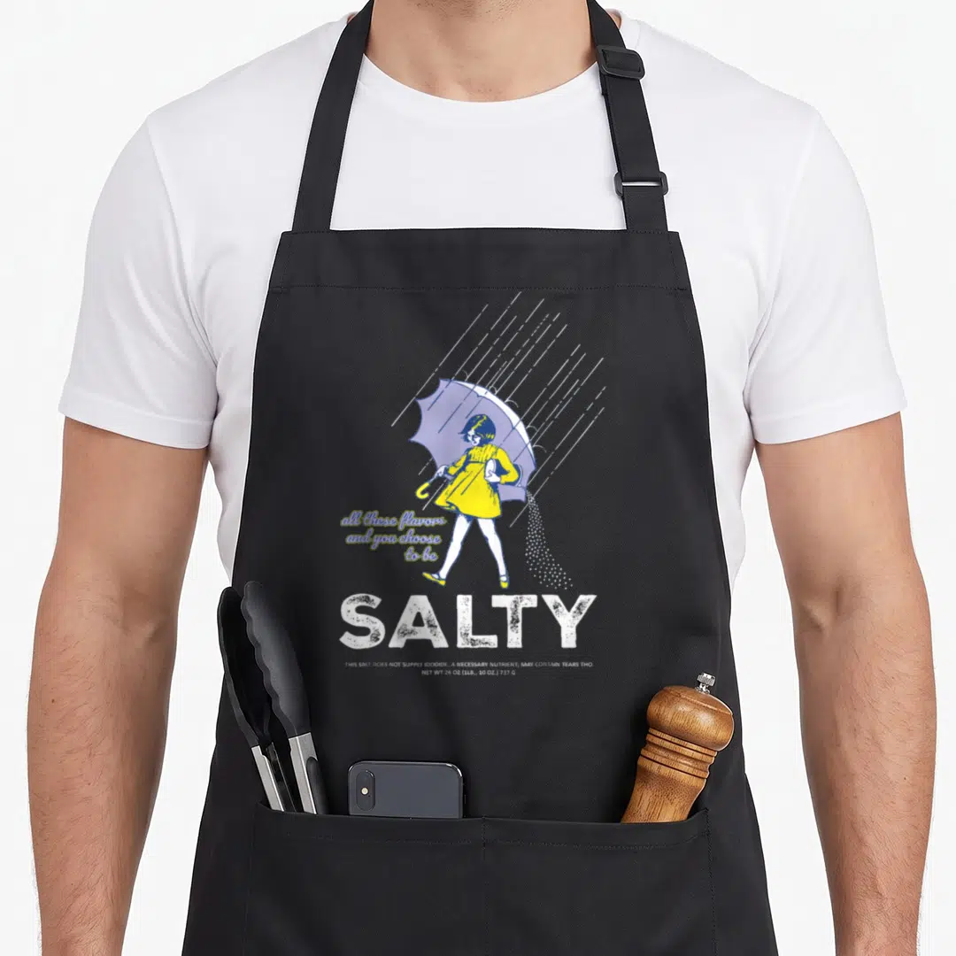 Salty sprinkle apron - Cartoon girl in yellow dress with purple umbrella pouring salt, white text reading SALTY — shown on a black cotton canvas apron against a white background