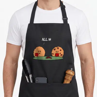 Chocolate cookie apron - Two brown cookies holding red cards at green table with ALL IN text and red chip pile — shown on a black cotton canvas apron against a white background