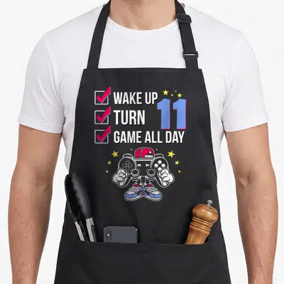 Gamer level birthday apron - WAKE UP TURN 11 GAME ALL DAY text with checklist, blue 11, and controller character — shown on a black cotton canvas apron against a white background