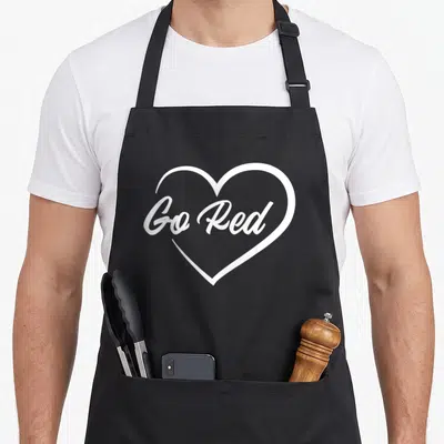 Month heart disease awareness apron - White heart outline containing Go Red cursive text on dark background — shown on a black cotton canvas apron against a white background