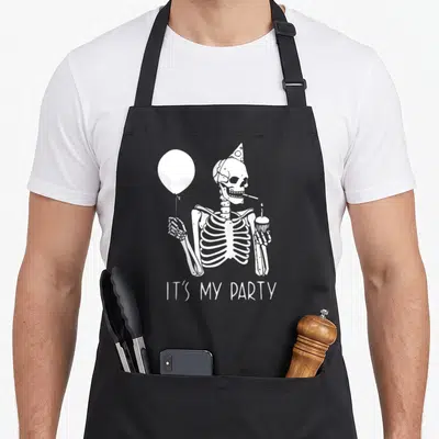 Costume funny halloween apron - White cartoon skeleton wearing a party hat holding a balloon and cupcake — shown on a black cotton canvas apron against a white background