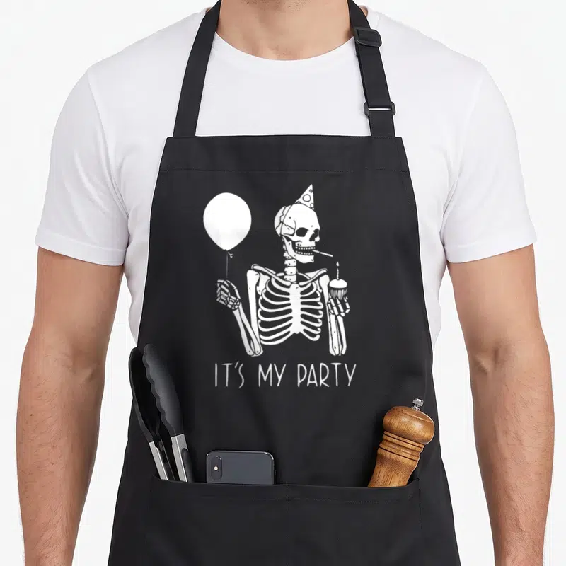 Costume funny halloween apron - White cartoon skeleton wearing a party hat holding a balloon and cupcake — shown on a black cotton canvas apron against a white background