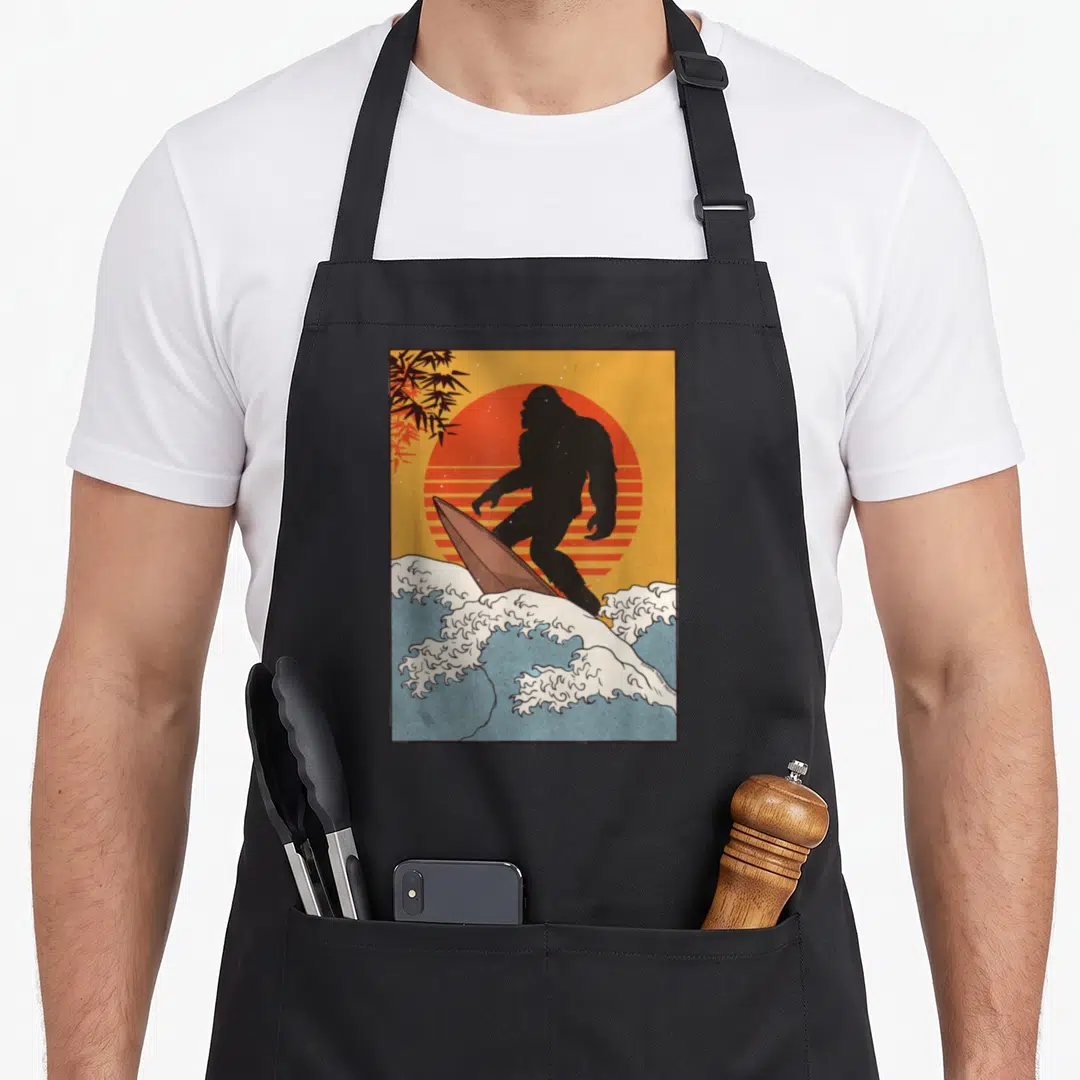 Kanagawa bigfoot apron - Black bigfoot surfing on blue waves against striped orange sun with bamboo leaves — shown on a black cotton canvas apron against a white background