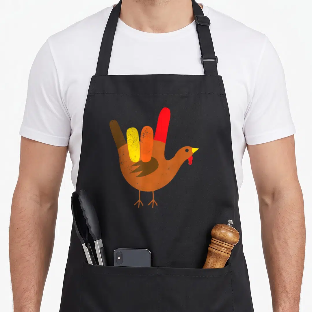 Sign thanksgiving apron - Cartoon turkey with four colored fingers forming ASL I love you sign in orange brown red — shown on a black cotton canvas apron against a white background