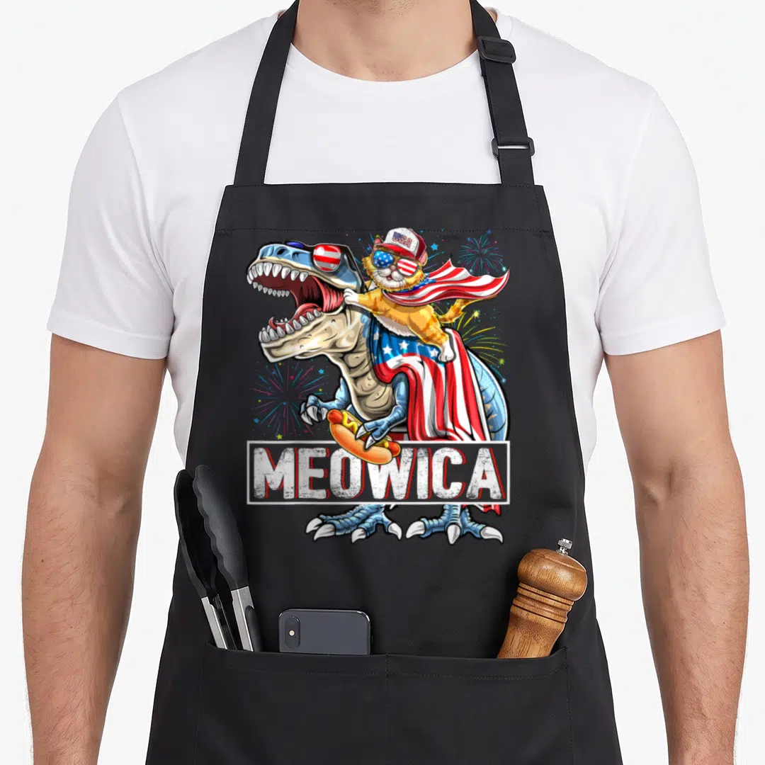 Rex 4th of july apron - Cat riding T-Rex in USA flag cape holding hot dog with MEOWICA text — shown on a black cotton canvas apron against a white background