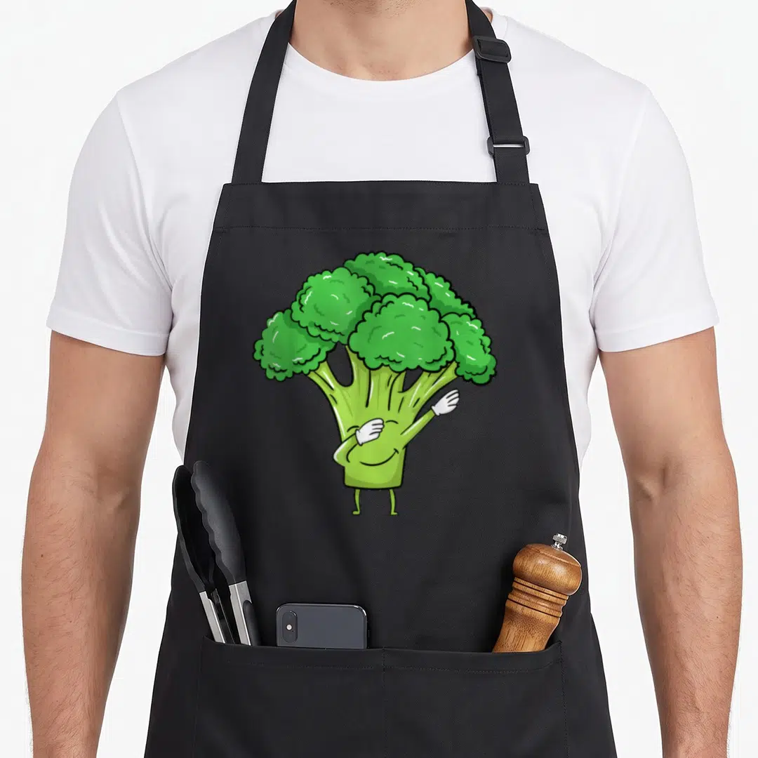 Dabbing broccoli vegetable apron - Green cartoon broccoli character with white gloved hands performing a dab pose — shown on a black cotton canvas apron against a white background
