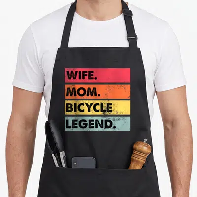 Bicycle mother's apron - Four horizontal bars in red orange yellow light blue with black text WIFE MOM BICYCLE LEGEND — shown on a black cotton canvas apron against a white background
