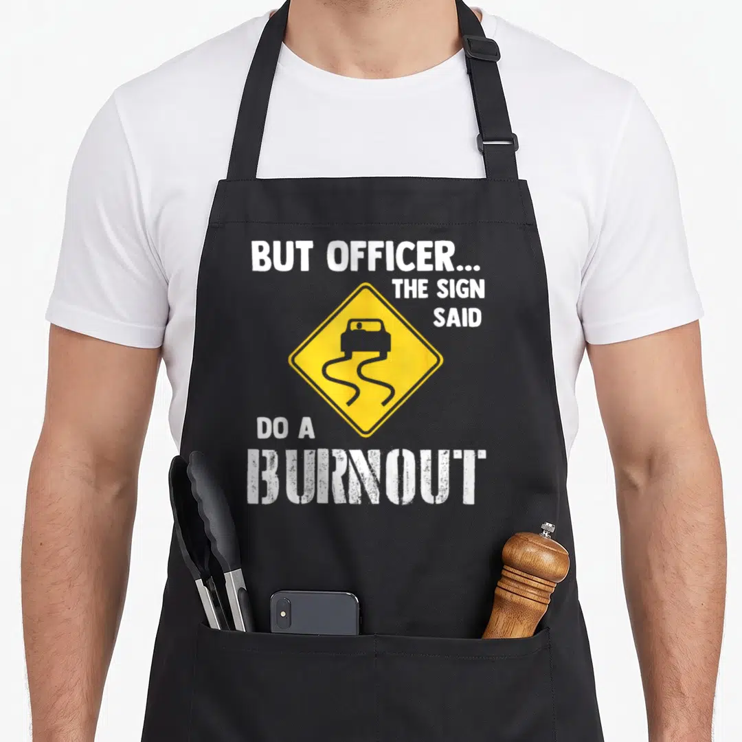 But car apron - Yellow diamond sign with car on wavy tracks and white burnout text — shown on a black cotton canvas apron against a white background