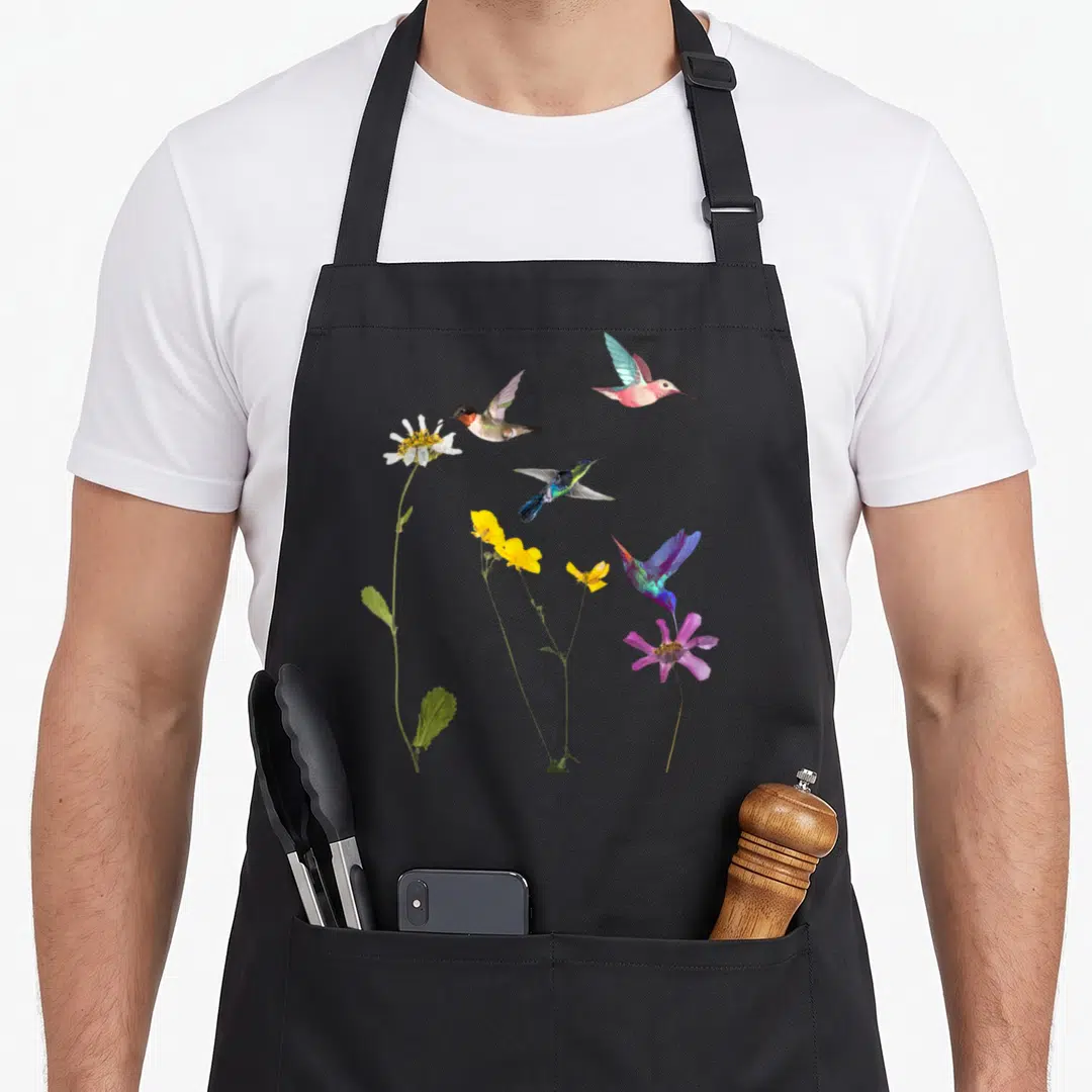 Flower hummingbird apron - Four hummingbirds in blue green pink yellow and white flying near three flowers on long stems — shown on a black cotton canvas apron against a white background