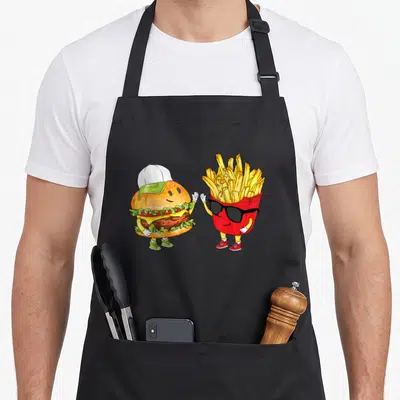 Hamburger fast food apron - Cartoon burger and fries high-fiving in yellow red green and brown — shown on a black cotton canvas apron against a white background