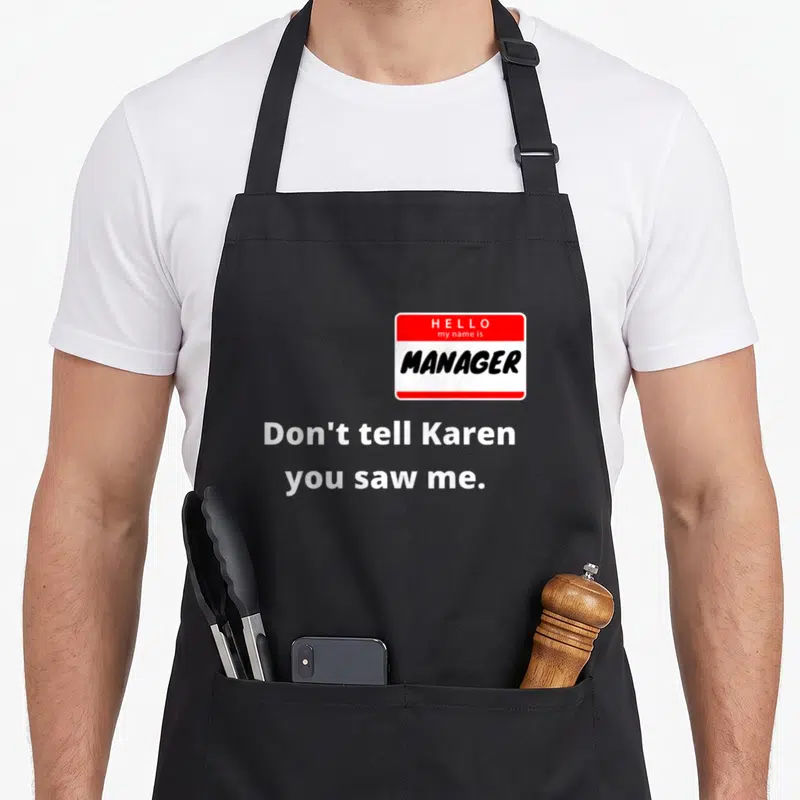 Hello couple apron - Red white name tag reading HELLO my name is MANAGER above white text Don't tell Karen you saw me — shown on a black cotton canvas apron against a white background