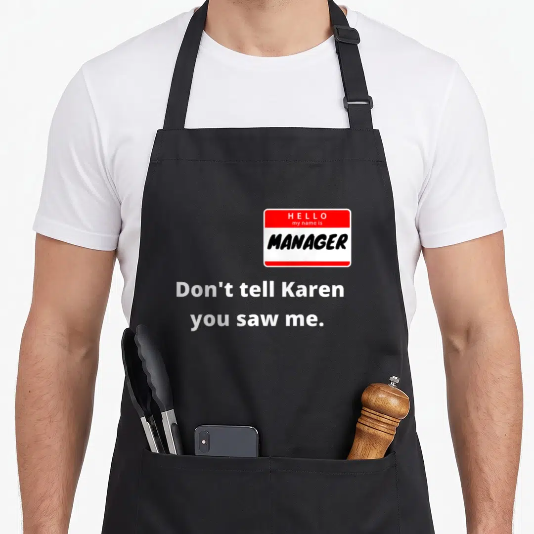 Hello couple apron - Red white name tag reading HELLO my name is MANAGER above white text Don't tell Karen you saw me — shown on a black cotton canvas apron against a white background