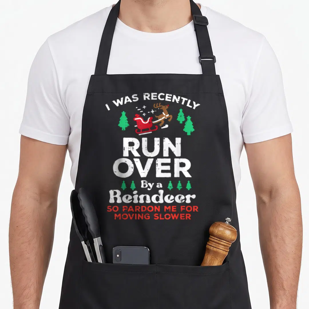 Runover reindeer apron - Cartoon reindeer pulling red sleigh between green trees with stacked white and red warning text — shown on a black cotton canvas apron against a white background