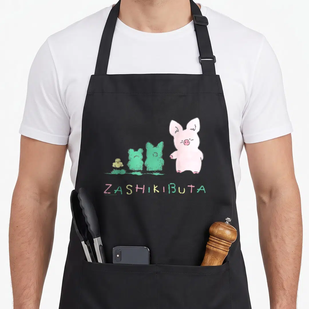 Zashikibuta general apron - Pink pig, two green bushes, yellow bird, and ZASHIKIBUTA text in cartoon style — shown on a black cotton canvas apron against a white background