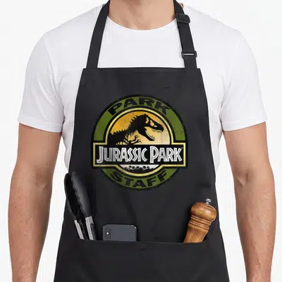 Staff distressed apron - Circular Jurassic Park Staff emblem with T-Rex silhouette in distressed green and yellow — shown on a black cotton canvas apron against a white background
