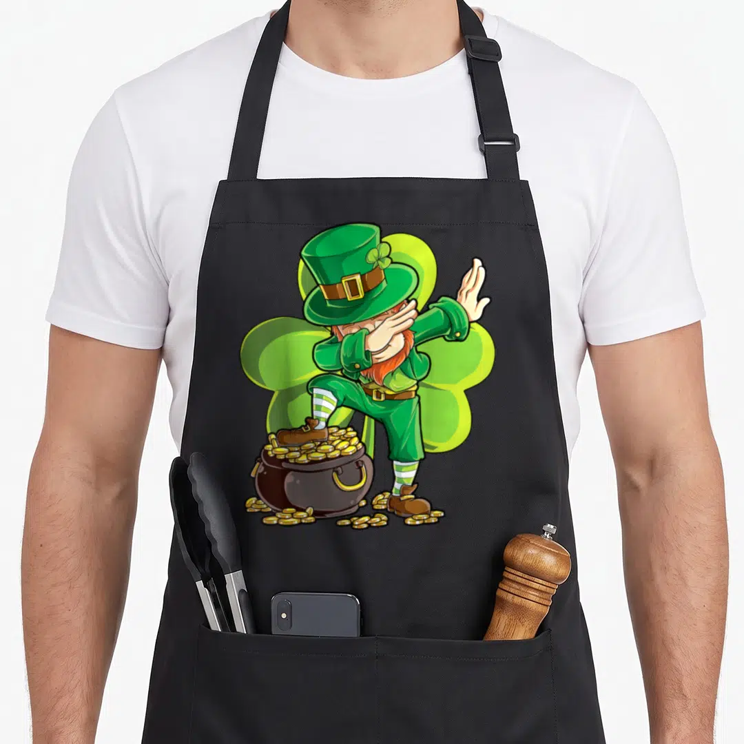 Dance st patrick day aprons - Cartoon leprechaun dabbing pose standing on a pot of gold coins in green and brown — shown on a black cotton canvas apron against a white background