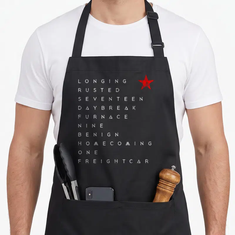 Trigger winter apron - Vertical list of ten white distressed words next to a single red star symbol — shown on a black cotton canvas apron against a white background
