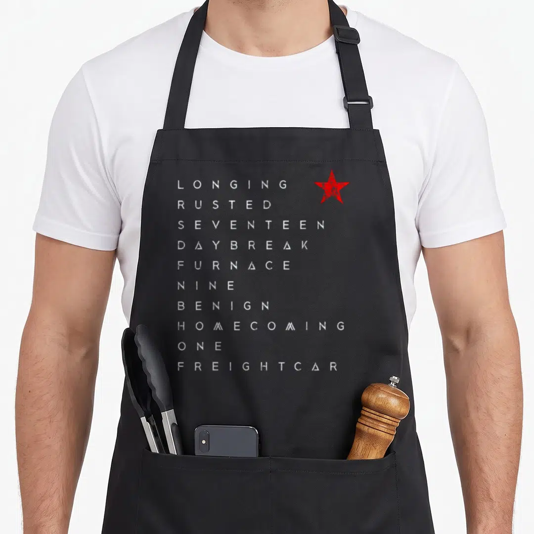 Trigger winter apron - Vertical list of ten white distressed words next to a single red star symbol — shown on a black cotton canvas apron against a white background